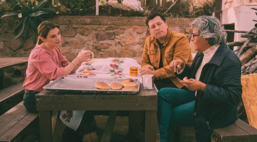 Image of activist and architect Ronald Rael wearing a brown jacket seated between Patti Jinich in a pink button down and blue jeans on the left and Angelica Macias in a black jacket with a white shirt and blue jeans on the right sharing fresh bread